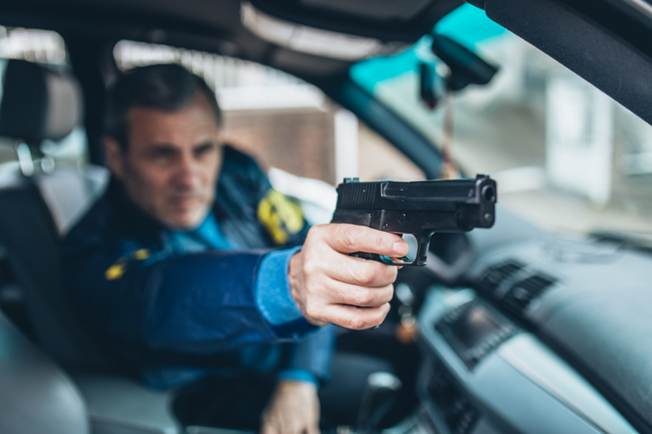 Middle aged man holding a gun in a car, wearing a dark blue jacket with yellow accents. The man's expression is serious and focused.